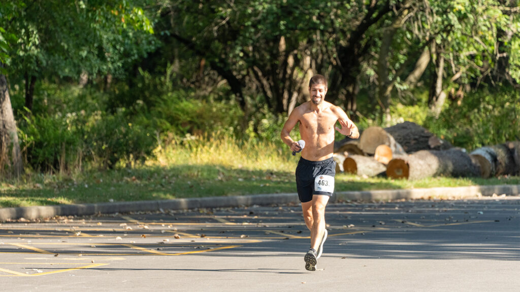 A shirtless man wearing black shorts and a race bib runs on a paved path near trees and large cut logs, smiling and holding a water bottle.