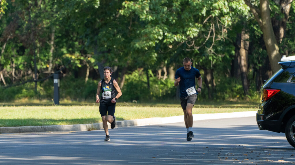 Two runners wearing race bibs compete on a paved road shaded by trees. The runner on the left wears black and the runner on the right wears blue. A black vehicle is parked on the right side, and greenery surrounds the area.