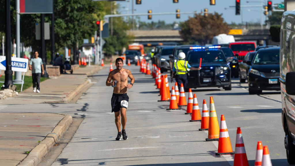 A shirtless male runner with a race bib jogs down a city street lined with orange traffic cones, with police cars, officers, and other vehicles in the background.