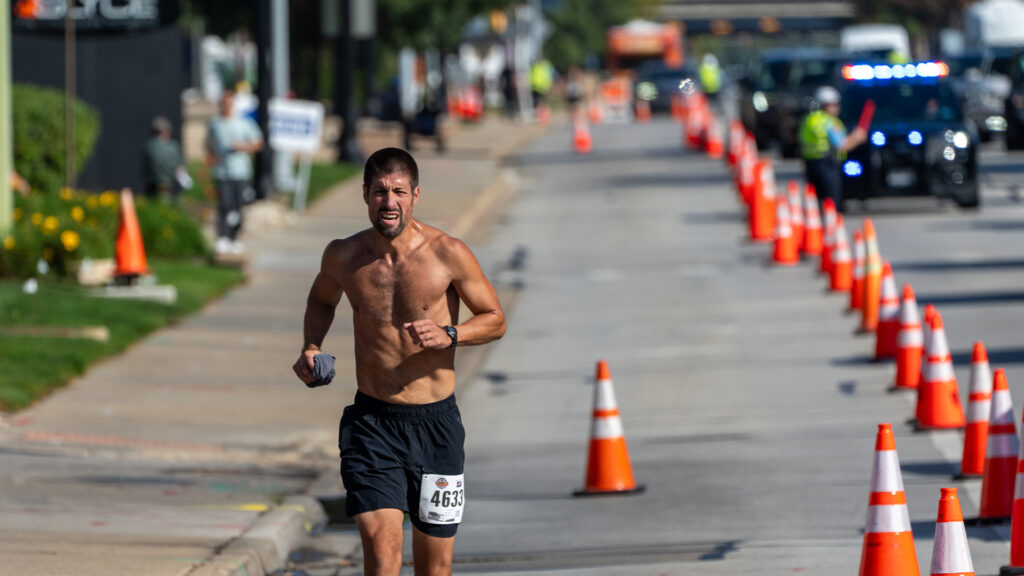 A shirtless male runner with a race bib and shorts runs on a city street lined with orange traffic cones during a race, with police cars and other people visible in the background.