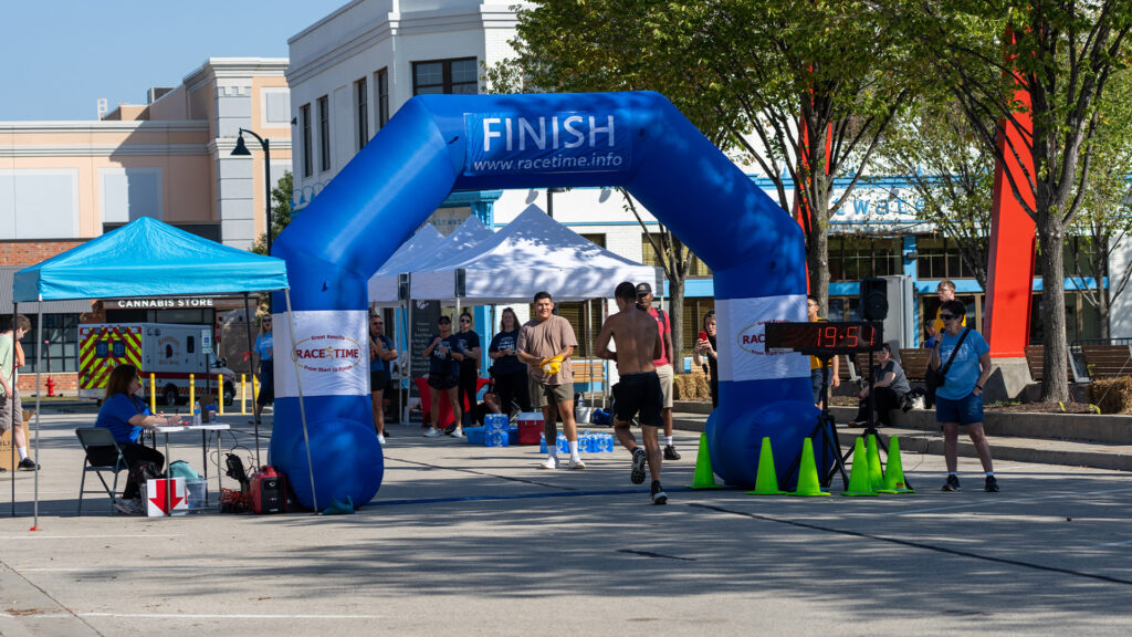 Runners approach a blue FINISH arch at a street race, with a digital clock showing 19:41. Spectators and staff stand nearby under tents. Trees and buildings line the street.