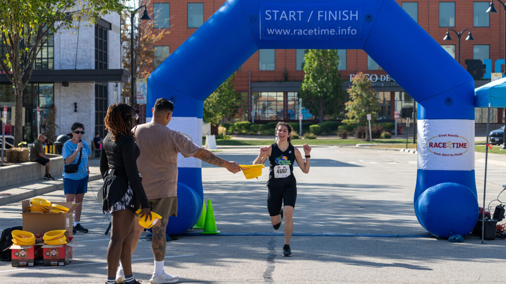A person in athletic gear is smiling and running toward the finish line under a blue inflatable arch as people cheer and hand out yellow items at a street race event.