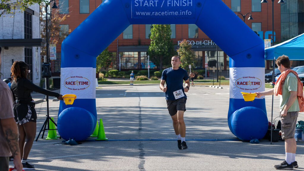 A runner in a blue shirt and black shorts crosses the finish line under a blue inflatable arch at a race event. People on the sides watch and cheer as he completes the race on a sunny day.