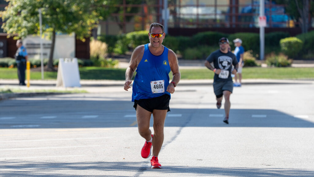 A man wearing a blue running tank top, black shorts, and red shoes runs in a road race, smiling and wearing sunglasses. Another runner follows behind on the street under clear daylight.