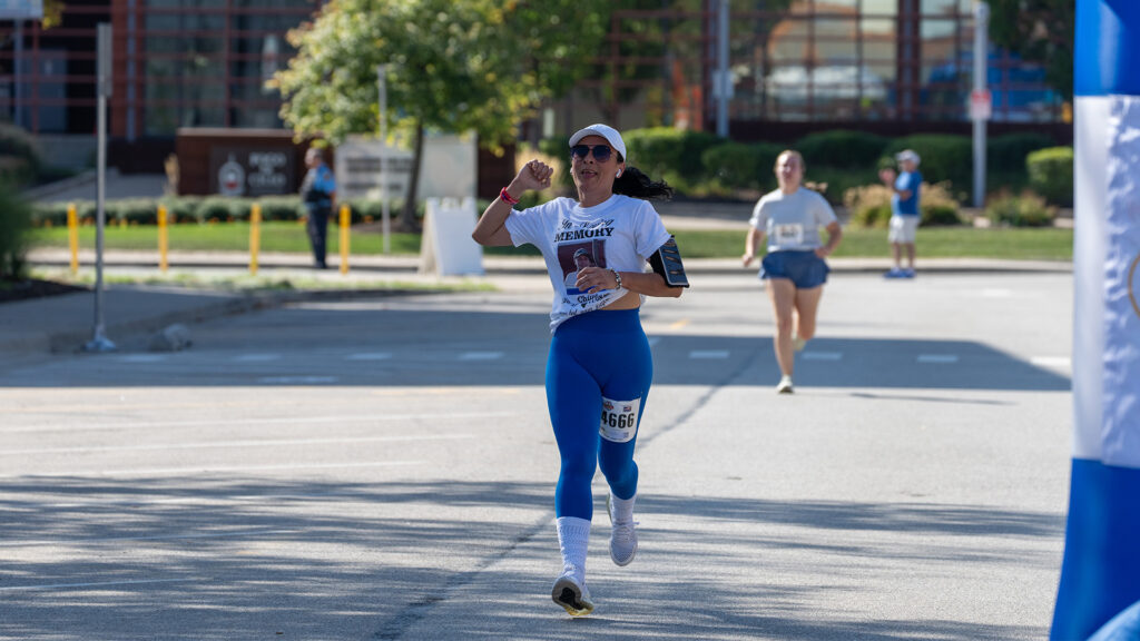 A woman in blue leggings and a white shirt runs toward the finish line in a road race, wearing sunglasses, a cap, and a race bib, with another runner following behind her on a sunny day.
