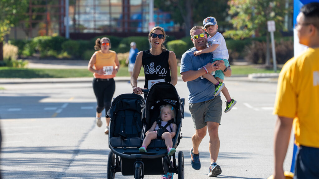 A smiling family participates in a race; a woman pushes a stroller with a young child, while a man carries another child. Other runners are visible in the background on a sunny day.
