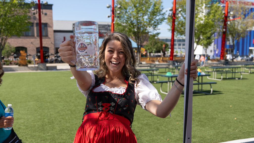 A smiling woman in a red and black traditional dress holds a large empty beer stein toward the camera at an outdoor event with picnic tables and trees in the background.