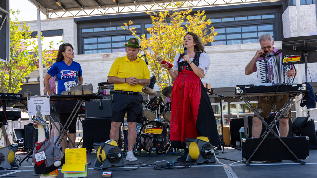 A band performs on an outdoor stage. Four adults stand among musical instruments: a woman in a blue shirt, a man in a yellow shirt and green hat, a woman in a red and black dress singing, and a man playing accordion.