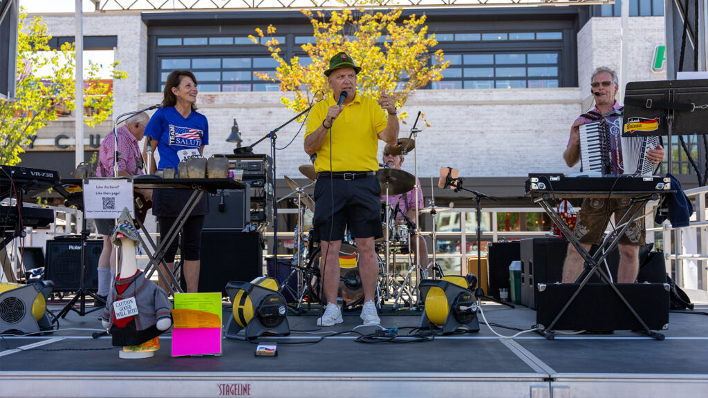 A band performs on an outdoor stage. The man in the center, wearing a yellow shirt and green hat, sings into a microphone. Other musicians play keyboard, accordion, and drums. Equipment and signs are visible around them.