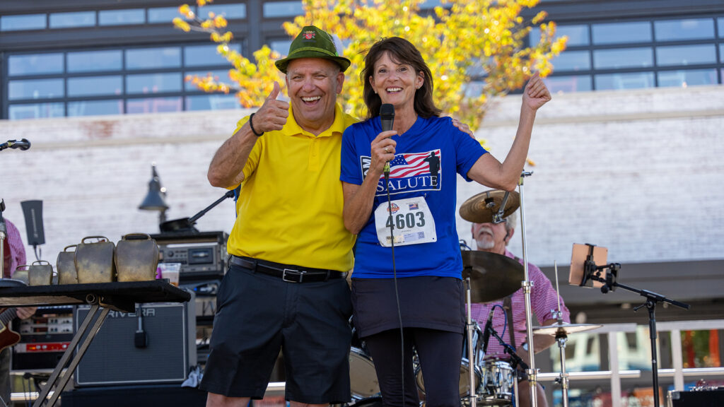 A smiling man in a yellow shirt and green hat gives a thumbs up while standing next to a woman with a microphone and Salute race bib. They are on an outdoor stage with musical instruments behind them.