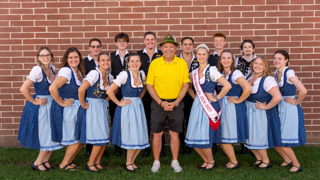 A group of smiling people in blue traditional folk costumes stands in a semicircle around a man in a yellow shirt and green hat, in front of a brick wall. One woman wears a sash reading Miss Bavarian Society 2022-2023.