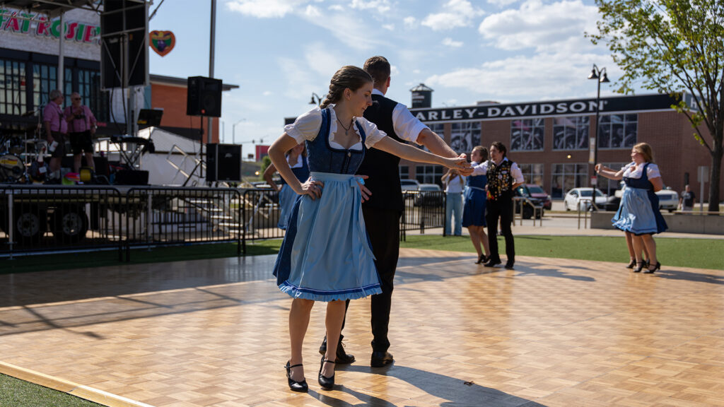 People in traditional Bavarian clothing dance on an outdoor wooden stage. A live band plays in the background. The event takes place in a sunny urban setting near Harley-Davidson signage.