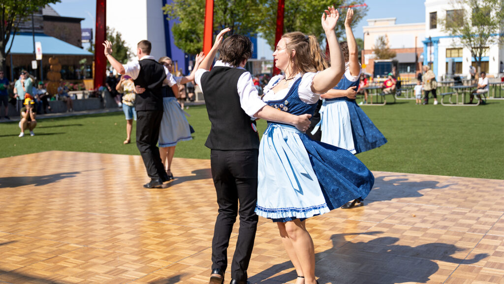 A group of people in traditional Bavarian attire dance on a wooden outdoor floor, with two pairs spinning and raising their arms. Onlookers sit on benches in the background on a sunny day.