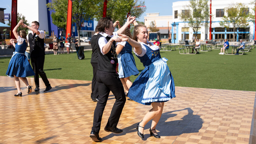 Four pairs of people in traditional Bavarian outfits dance on a wooden outdoor stage on a sunny day, with green grass and buildings in the background.