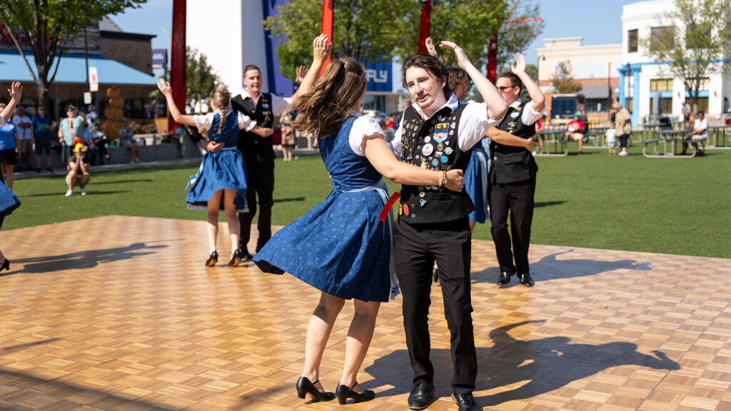 A group of people in traditional attire are dancing on a wooden outdoor stage in a park, with trees and buildings in the background under a sunny sky.