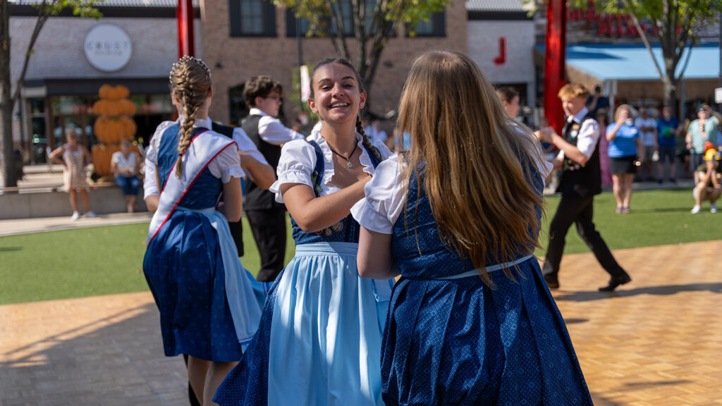Young women and men in traditional blue and white outfits dance outdoors on a sunny day, with a crowd of people watching in the background. The dancers are smiling and holding hands.