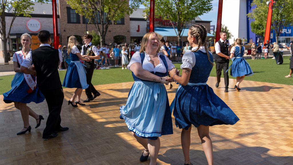 People dressed in traditional Bavarian attire dance on an outdoor wooden floor during a sunny festival, with onlookers and buildings in the background.