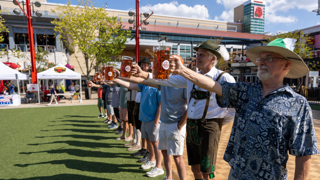 A line of people outdoors each hold out a large glass stein of beer in one hand during an Oktoberfest event, with festival tents and buildings visible in the background.