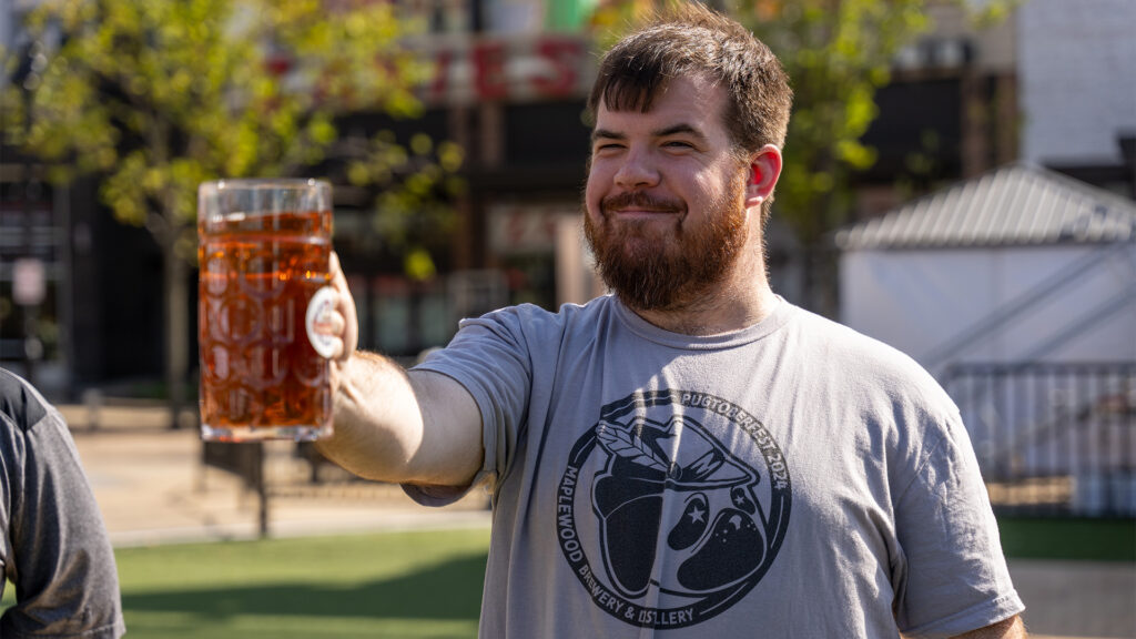 A bearded man with short hair smiles while holding a large glass mug of amber beer outdoors. He is wearing a light gray T-shirt with a graphic design, standing in a sunny area with trees and buildings in the background.