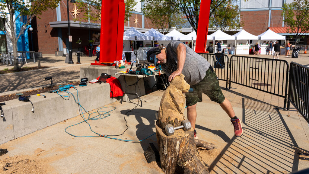 A person wearing safety gear carves a wooden sculpture outdoors with power tools, surrounded by wood shavings, barriers, and tents on a sunny day.