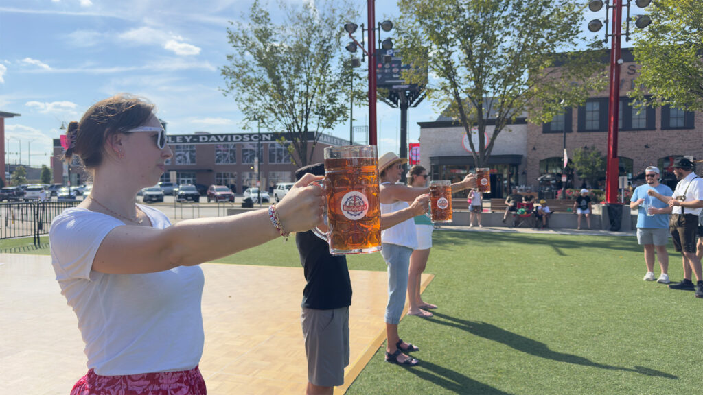 Three people stand outdoors on a sunny day, each holding a large mug of beer with an outstretched arm, participating in a stein-holding contest. There are buildings and trees in the background.