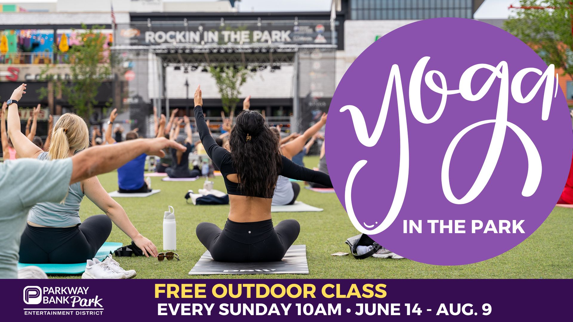 People participate in an outdoor yoga class on a lawn, with a large sign reading Yoga in the Park and event details displayed.