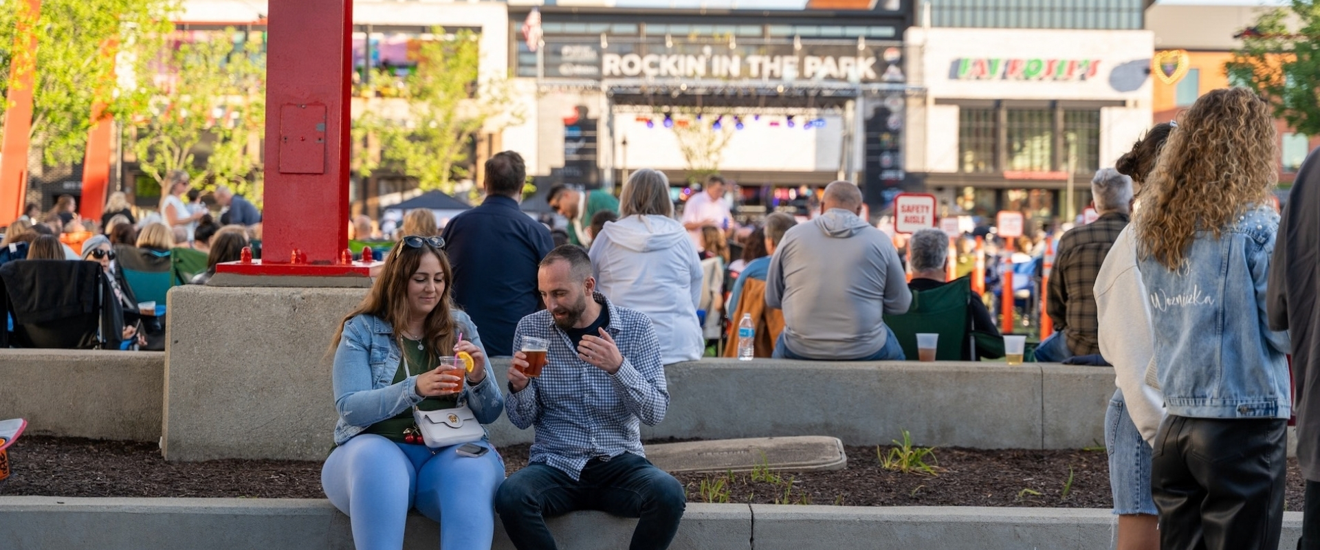 A man and a woman sit on a stone wall drinking from plastic cups in front of an outdoor concert stage.