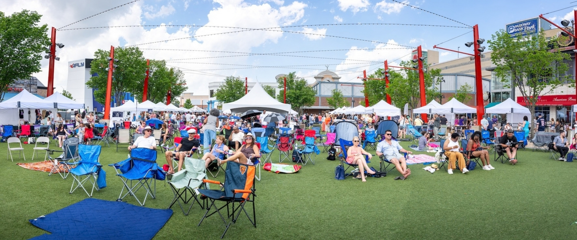 People relaxing in folding chairs on a lawn at an outdoor event surrounded by white vendor tents.