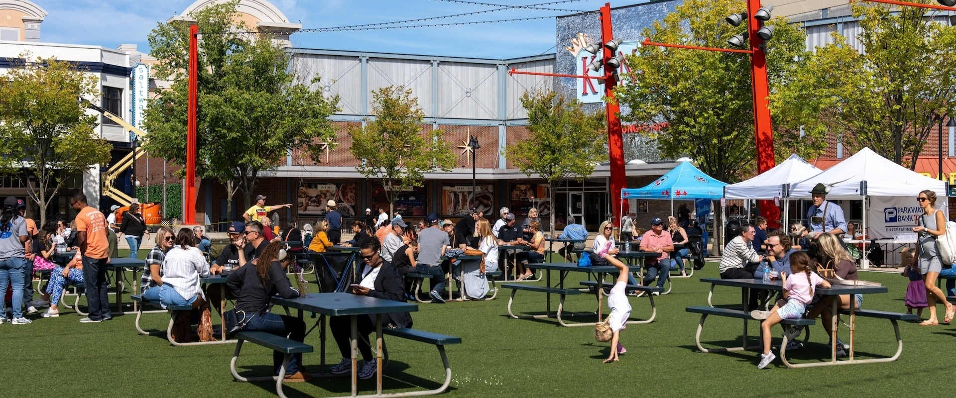 People sitting at outdoor tables in a shopping plaza with white canopy tents and a child doing a handstand on the grass.