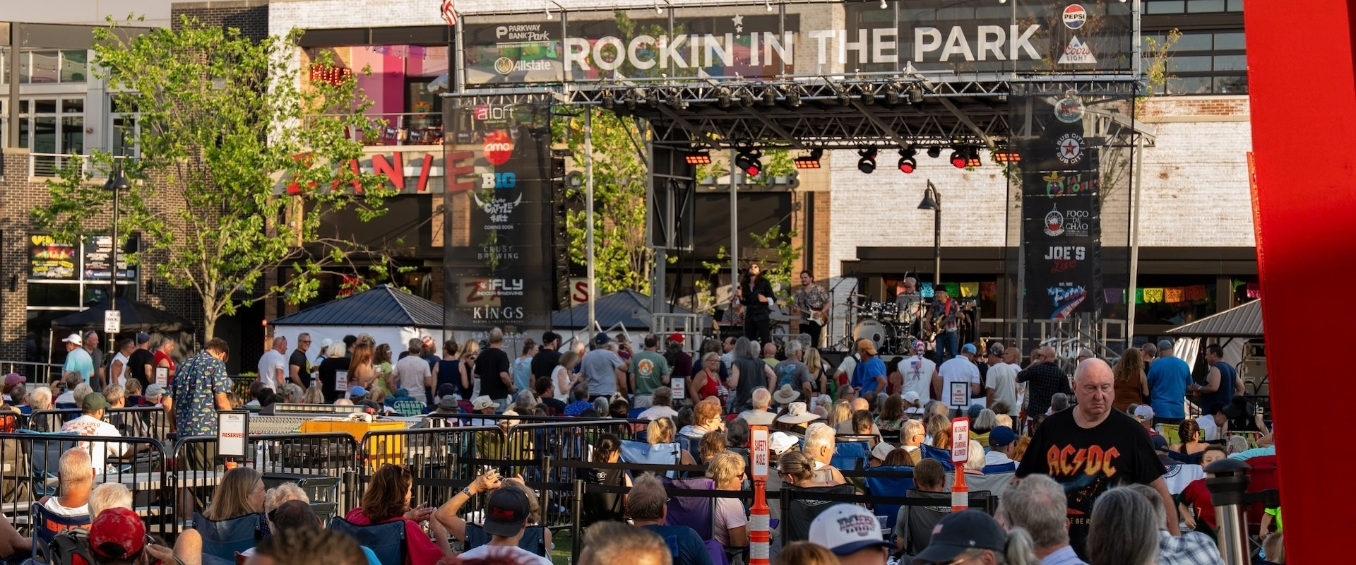 A crowd sits and stands in an outdoor plaza watching a band perform on a stage under a banner that reads Rockin in the Park.