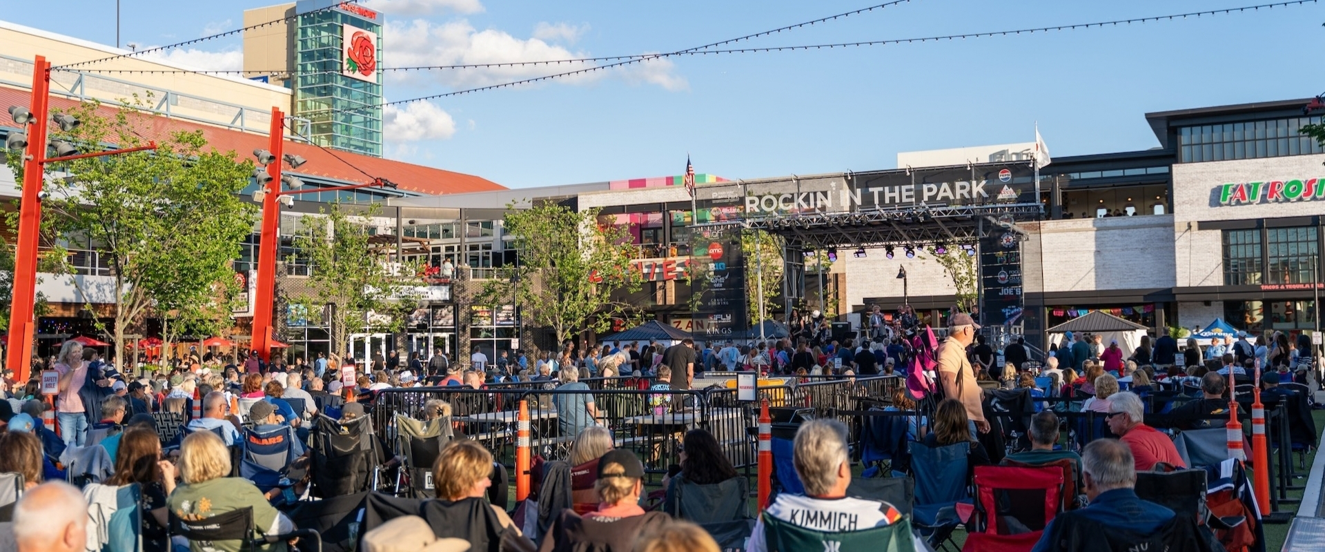A large crowd sits in folding chairs at an outdoor concert venue in front of a stage labeled Rockin in the Park.