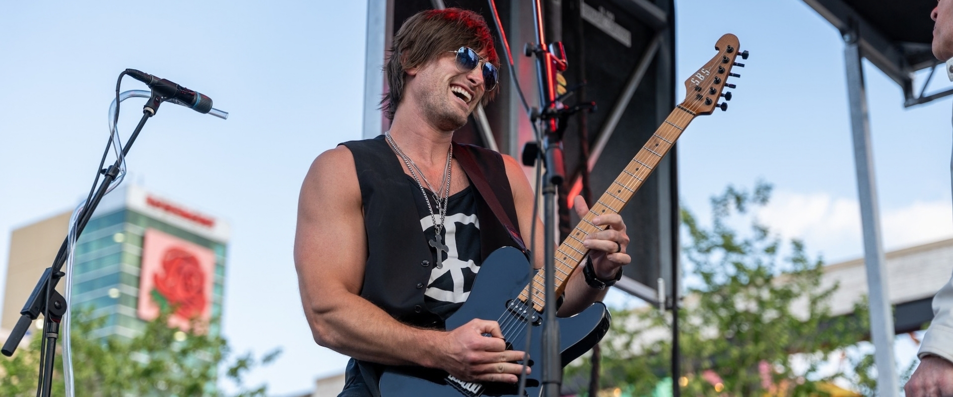 A smiling musician with sunglasses playing an electric guitar on an outdoor stage.