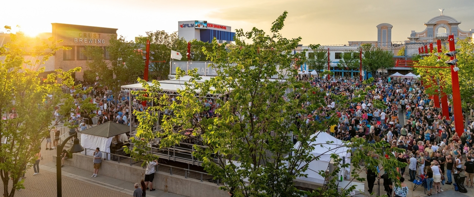A large crowd gathers at an outdoor music venue during sunset, with a band playing on a central stage.