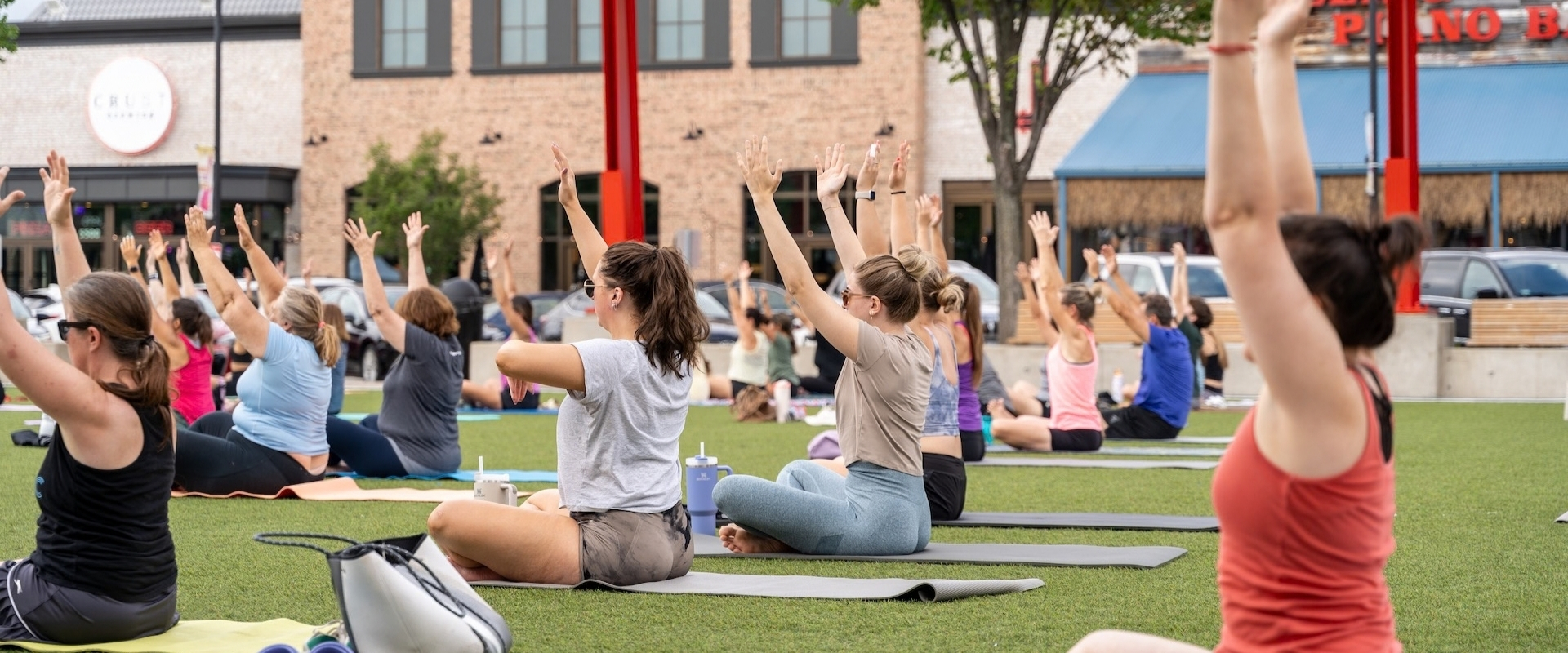 A group of people practicing outdoor yoga on mats on a lawn, with arms raised.